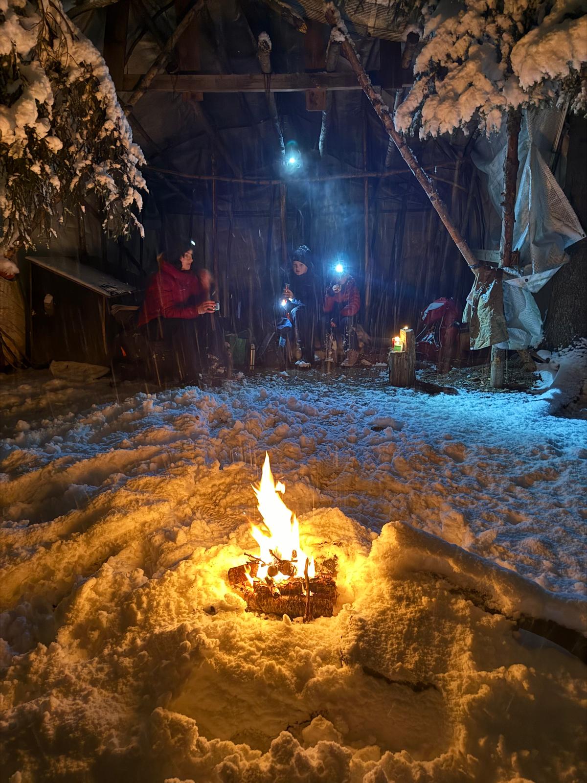 To personer sitter på reinskinn foran bålet i snøen i Kjønstadmarka